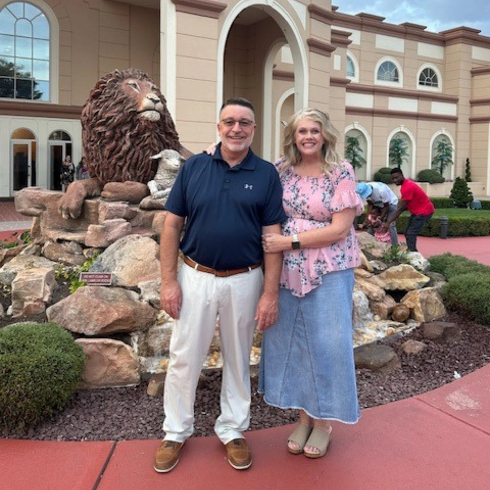 A man and a woman posing in front of a large lion statue and stone landscaping in front of a resort-style building.